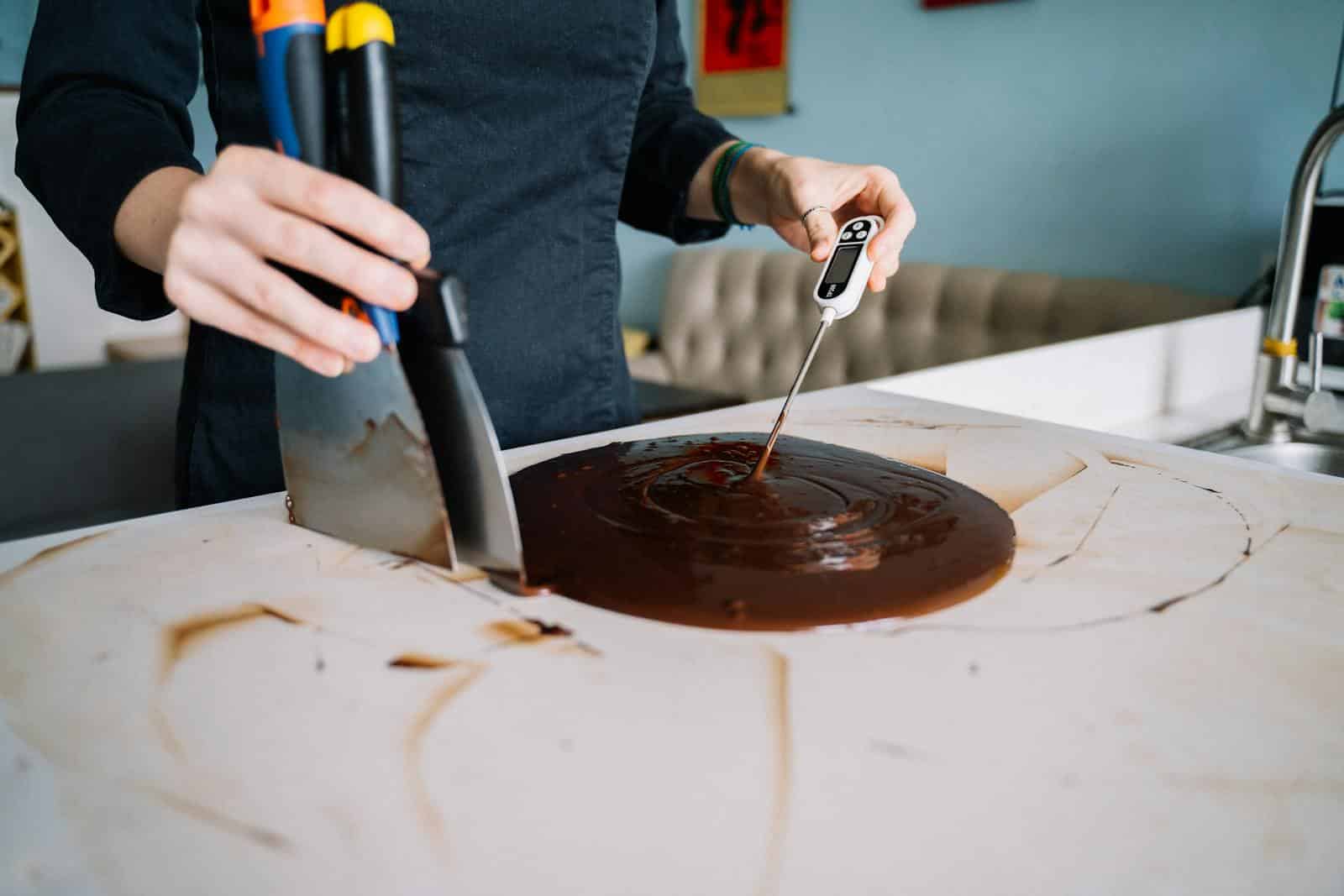 Close-up of a person tempering chocolate on a kitchen counter using a scraper and thermometer.